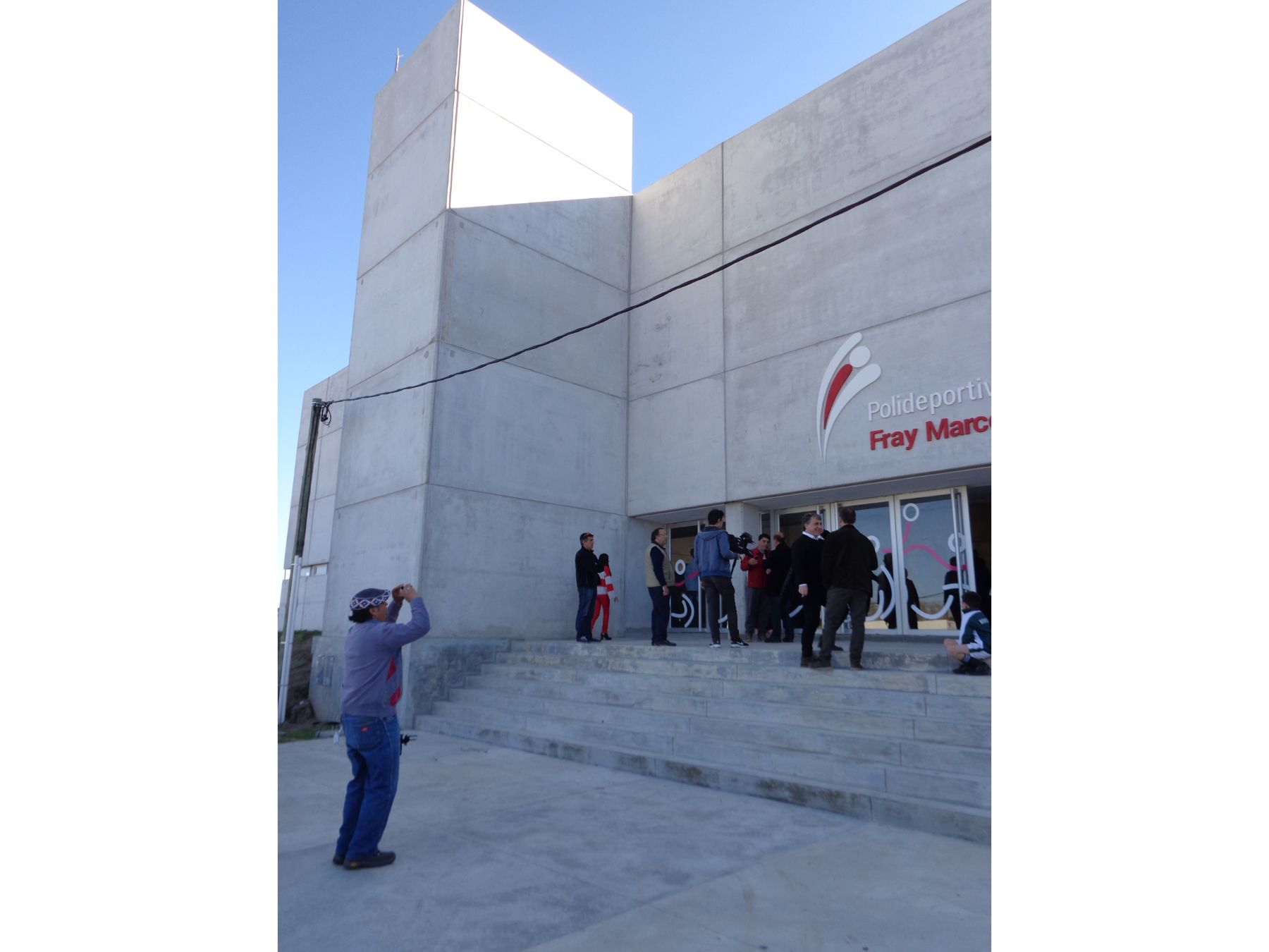 Es una fotografía de la fachada principal del polideportivo de Fray Marcos durante su inauguración. Es un edificio de hormigón de aproximadamente 5 metros de altura. Se aprecia la escalera principal con el logo sobre las puertas de entrada y un grupo de personas que acudieron a la inauguración.